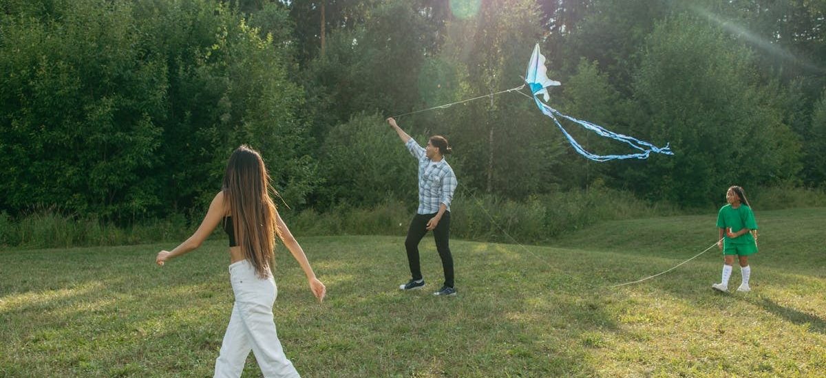 A family enjoys a sunny day flying a kite in a green park, surrounded by trees.